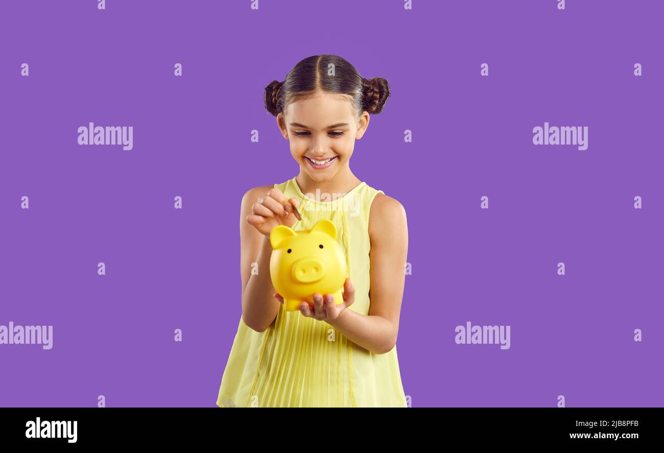 Smiling smart preteen girl throws coin into piggy bank isolated on purple background. Stock Photo