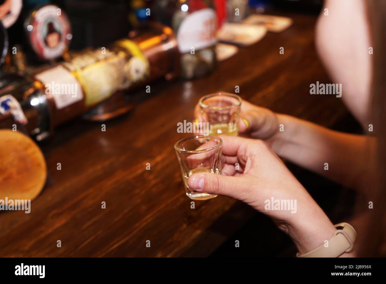 Two female hands clink glasses with alcoholic shots. Blurred bar background. Local cozy bar. Background for menu and alcohol card Stock Photo