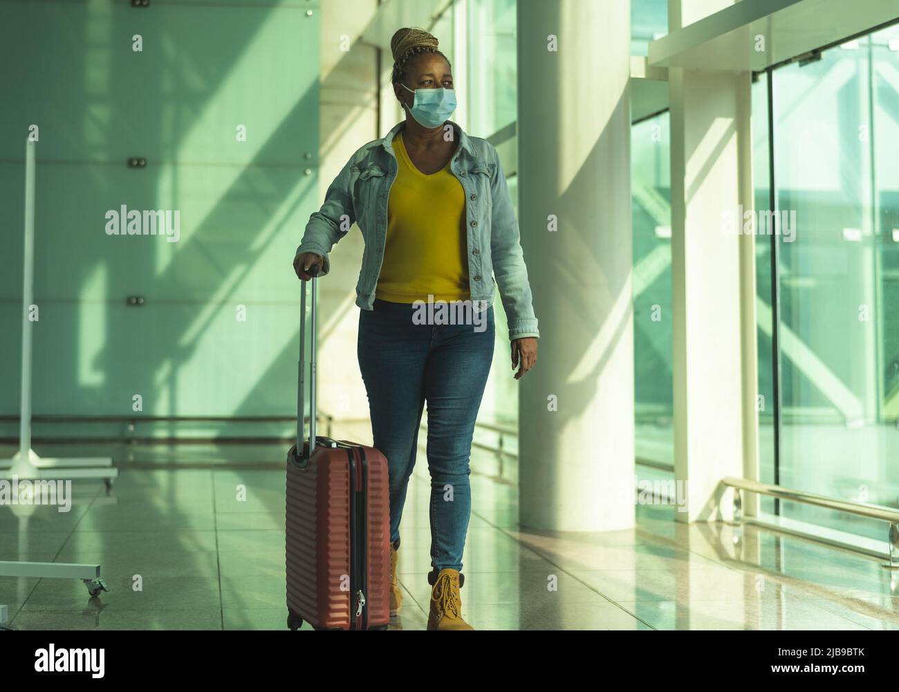 African senior woman walking in airport terminal with luggage wearing face mask during coronavirus pandemic Stock Photo