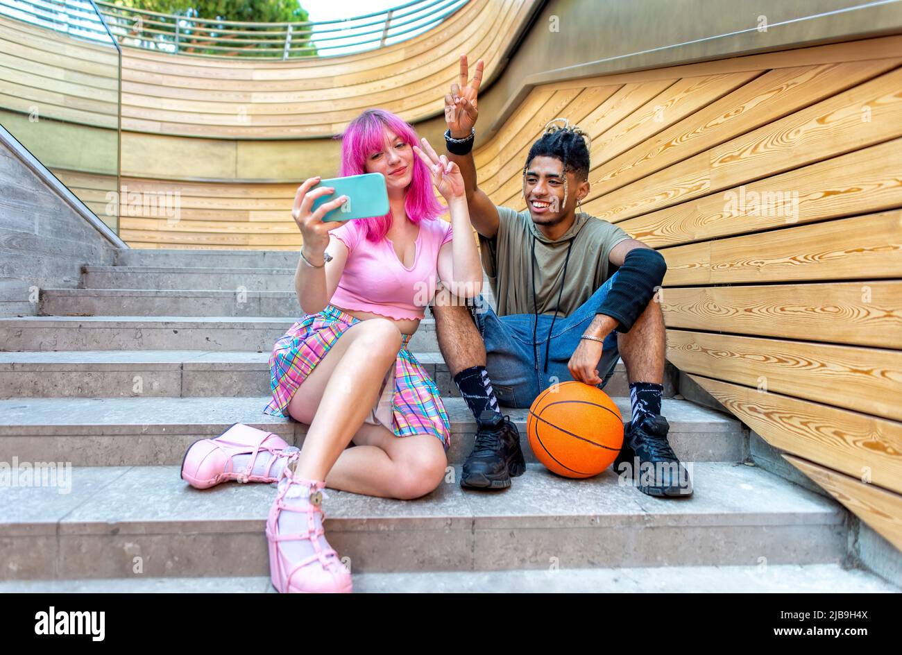 alternative diverse couple hanging out together sitting on city stairs making a selfie holding smartphone. interracial teenagers friends with ball Stock Photo