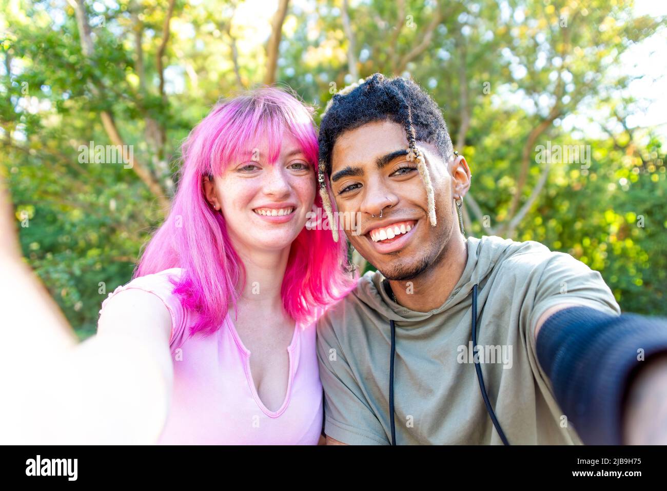 alternative diverse couple hanging out in a park making a pov selfie portrait holding smartphone on both side. interracial teenagers friends relaxing Stock Photo