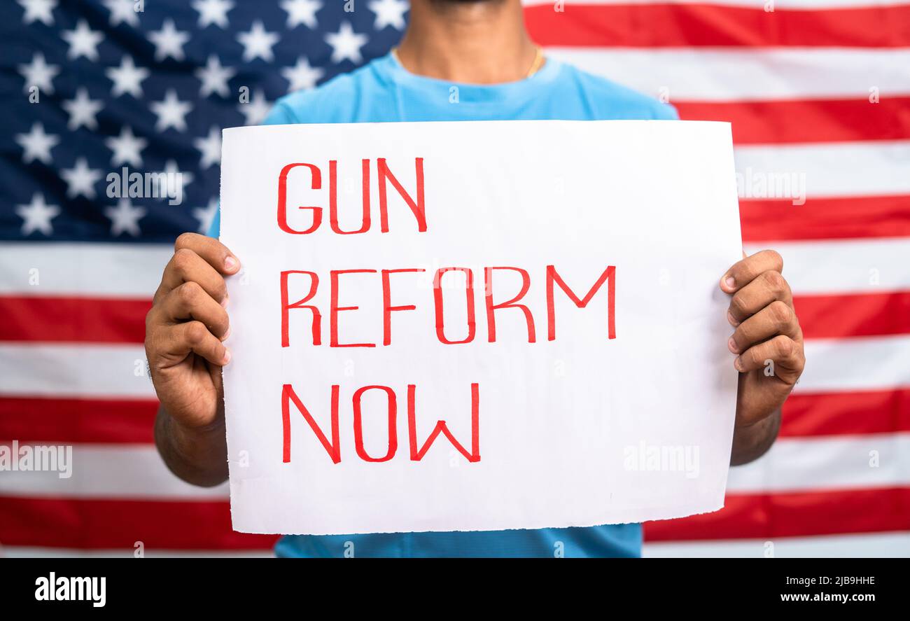 young man showing Gun reform Now sign board in front of US american flag - concept of protest against gun violence and activism. Stock Photo
