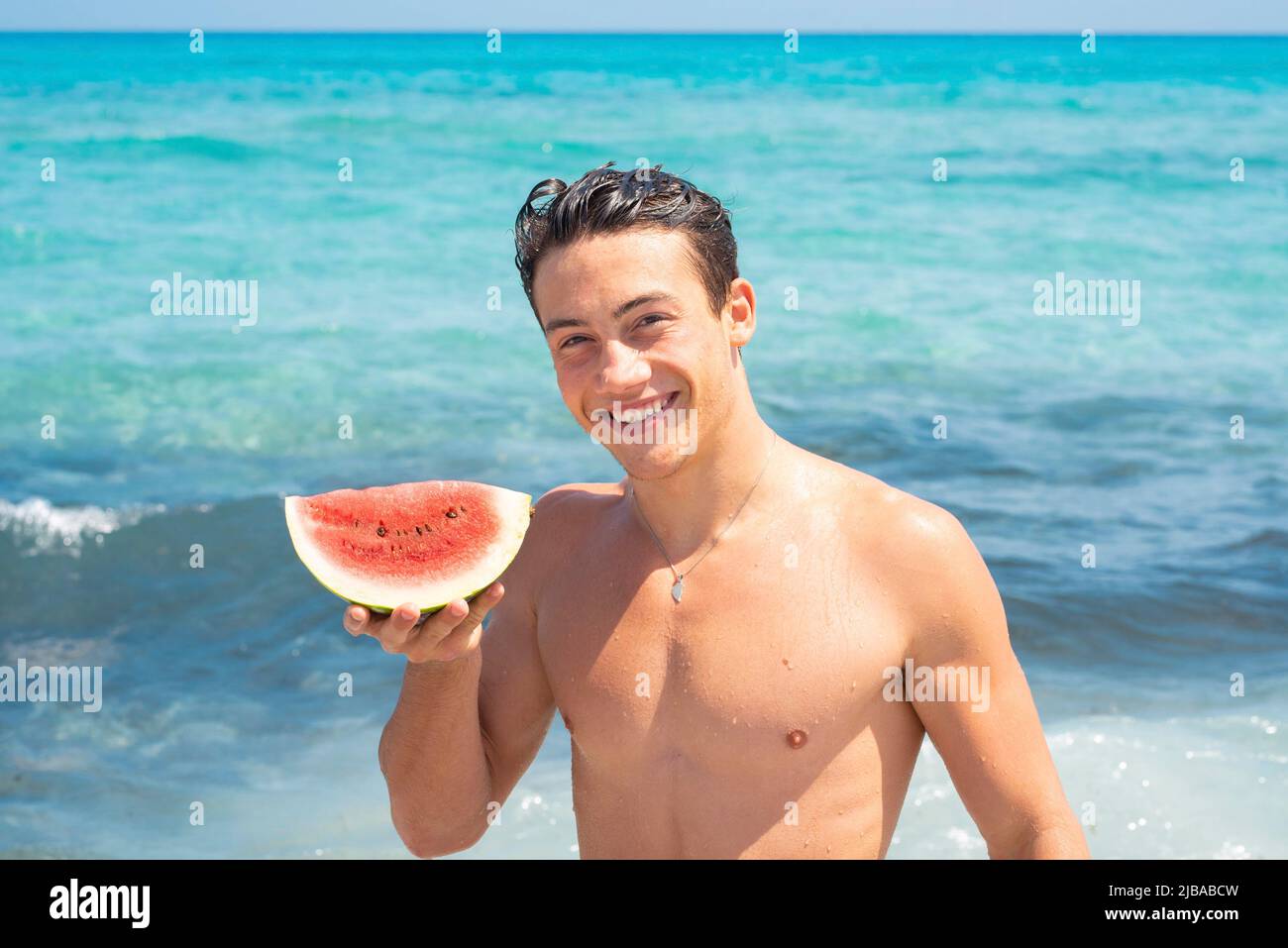 Young fit body boy portrait holding red fresh watermelon with blue ocean water in background. Summer holiday vacation season concept lifestyle people Stock Photo