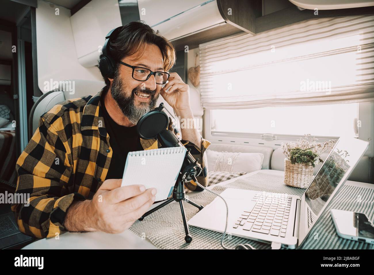 Modern adult man wokring with laptop and microphone inside a camper van in digital nomad job lifestyle. Technology and travel. Alternative freedom off Stock Photo