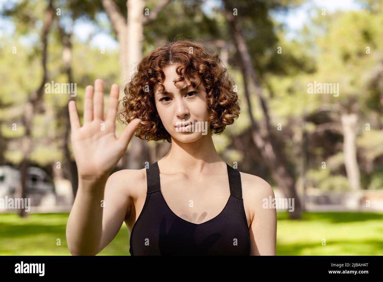Portrait of young redhead woman wearing sports bra standing on city park, outdoor doing stop sing with palm of the hand. Warning expression with negat Stock Photo