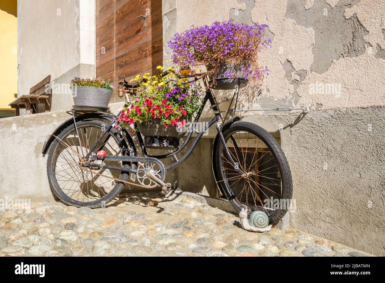 An old rusty bike, decorated with beautiful flowers, in the historic village center of S-Chanf (Upper Engadine Valley , Grisons, Switzerland) Stock Photo