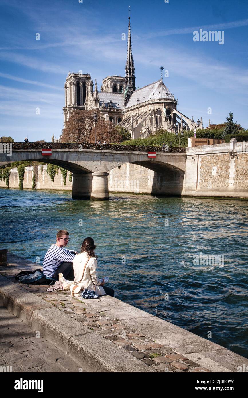 A couple enjoys the ideal weather along the River Seine below the Pont Archeveche bridge and Notre Dame in Paris, France. Stock Photo