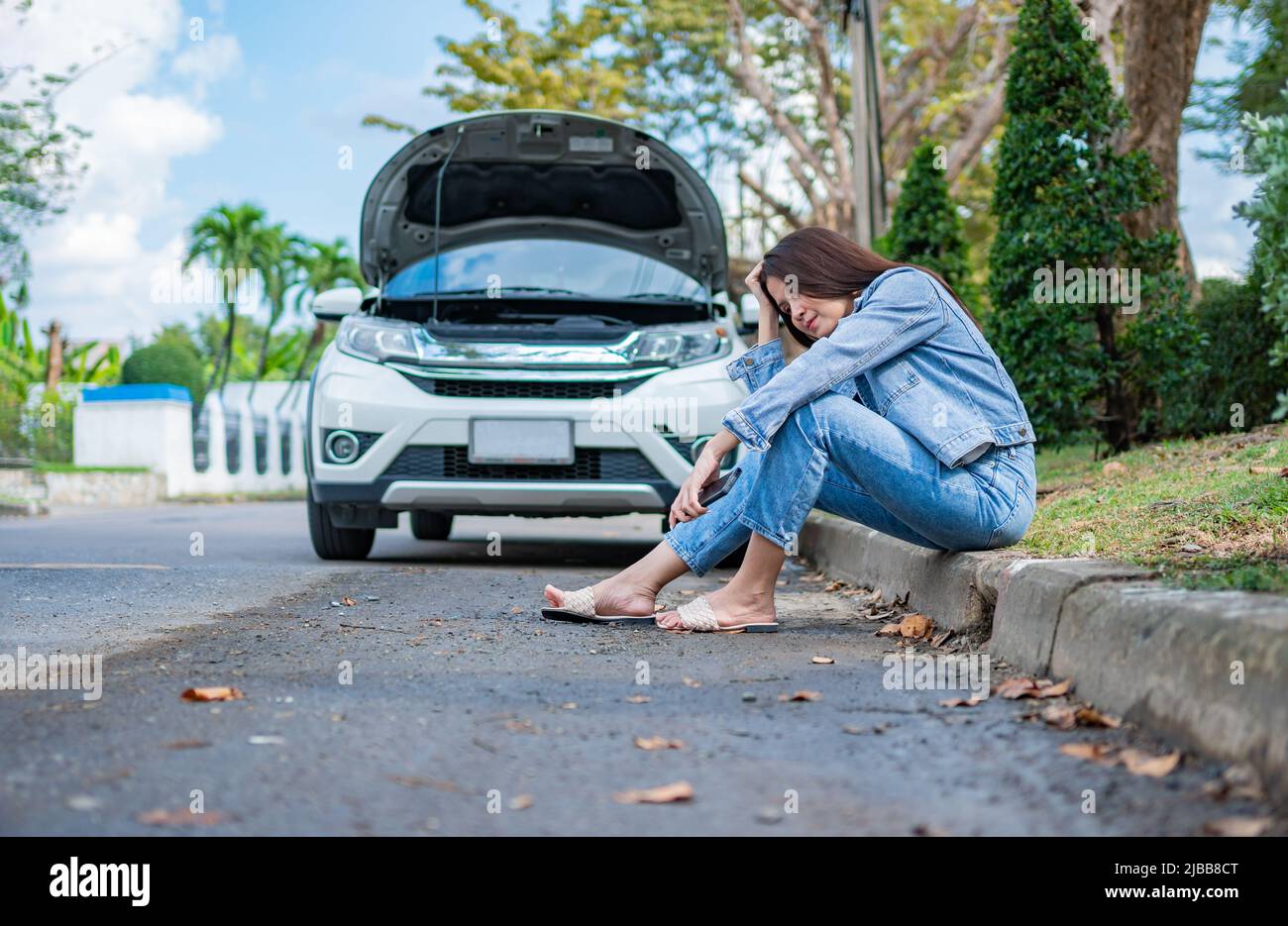 Asian woman sitting beside car after a car breakdown on street. Concept of vehicle engine problem or accident and emergency help from Professional mec Stock Photo
