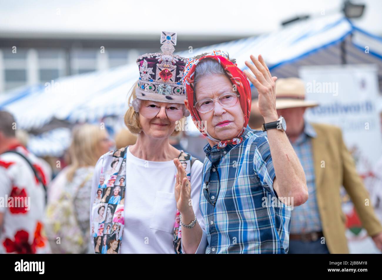 Two revellers wearing cardboard masks of the Queen. Platinum Jubilee celebrations in Kenilworth, Warwickshire. Thousands of locals enjoyed the main road closure, to sit on picnic tables and celebrate the Queen's 70th anniversary of her reign. Stock Photo