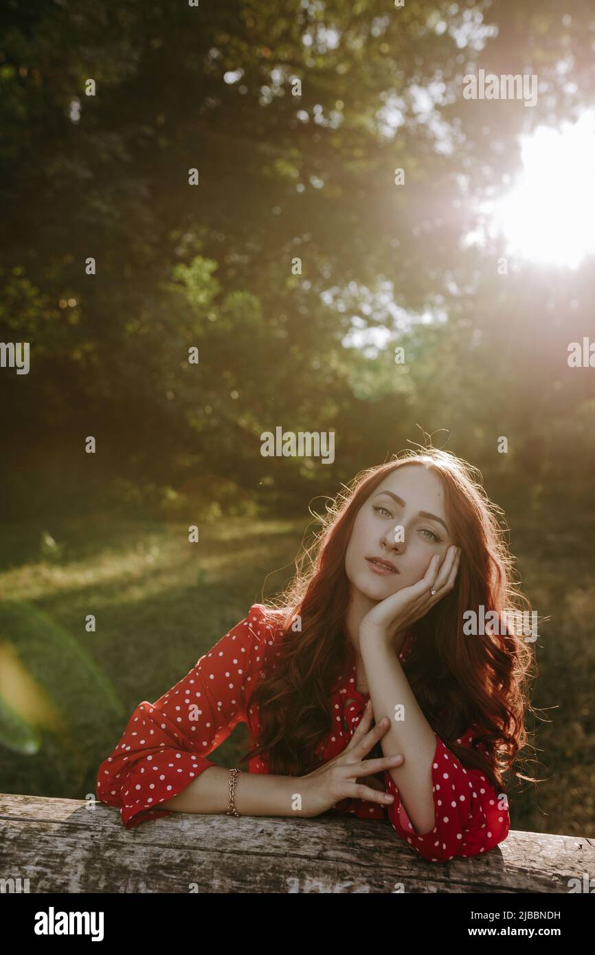 Portrait of pretty redhead woman in red summer loose shirt leaning by hands on a dry beam in wood and posing on a camera Stock Photo