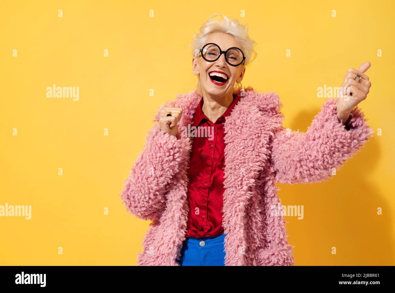 Happy and playful mature woman dancing, smiling and having fun. Photo of elderly woman above 70 years old in stylish outfit on yellow background Stock Photo