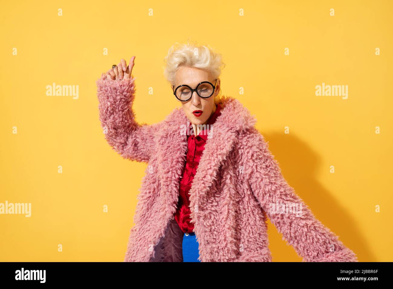 Happy and playful mature woman dancing, smiling and having fun. Photo of elderly woman above 70 years old in stylish outfit on yellow background Stock Photo