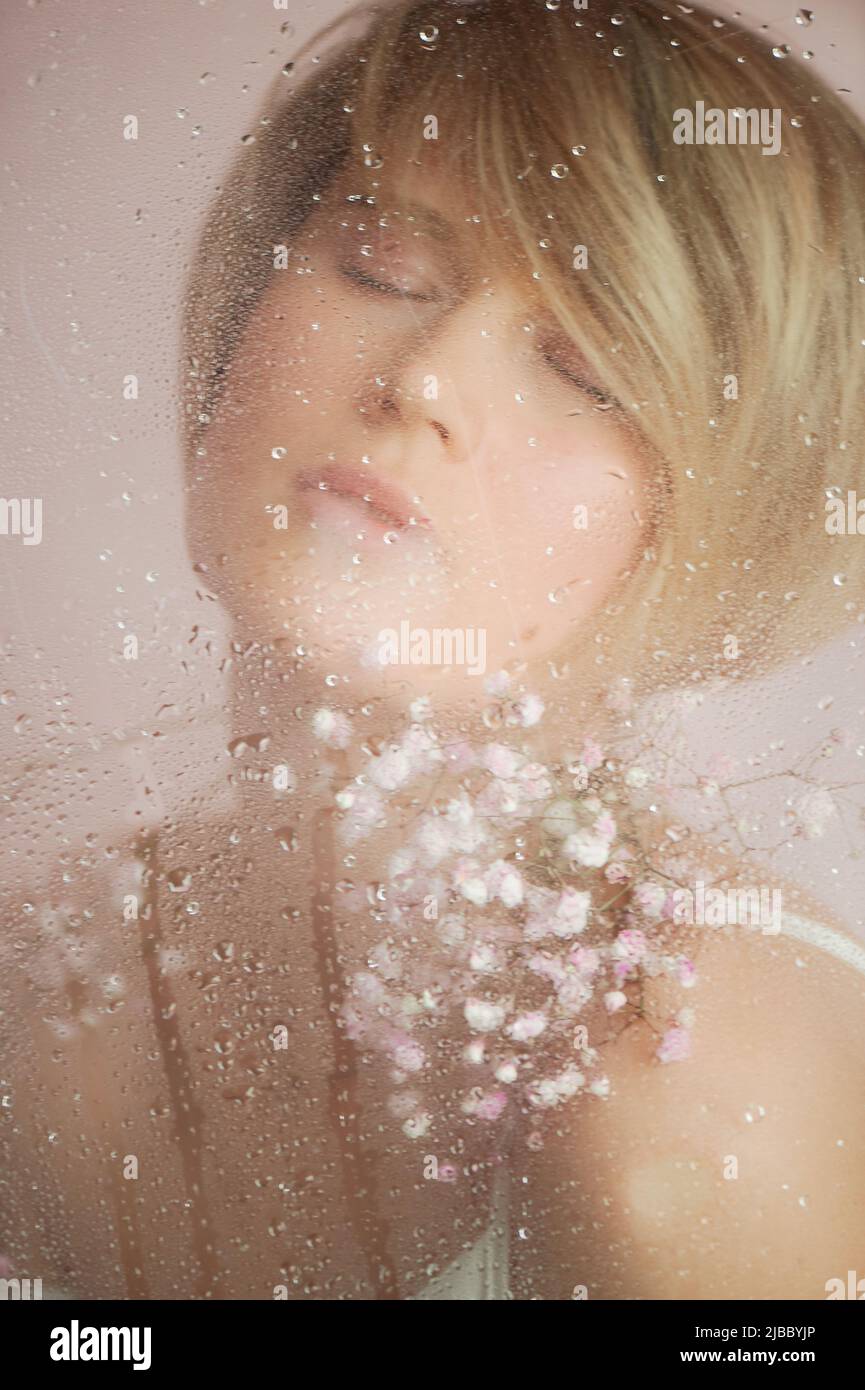 A girl with flowers behind wet glass. Nega and enjoyment of life. Eyes closed. Beauty and personal care industry Stock Photo