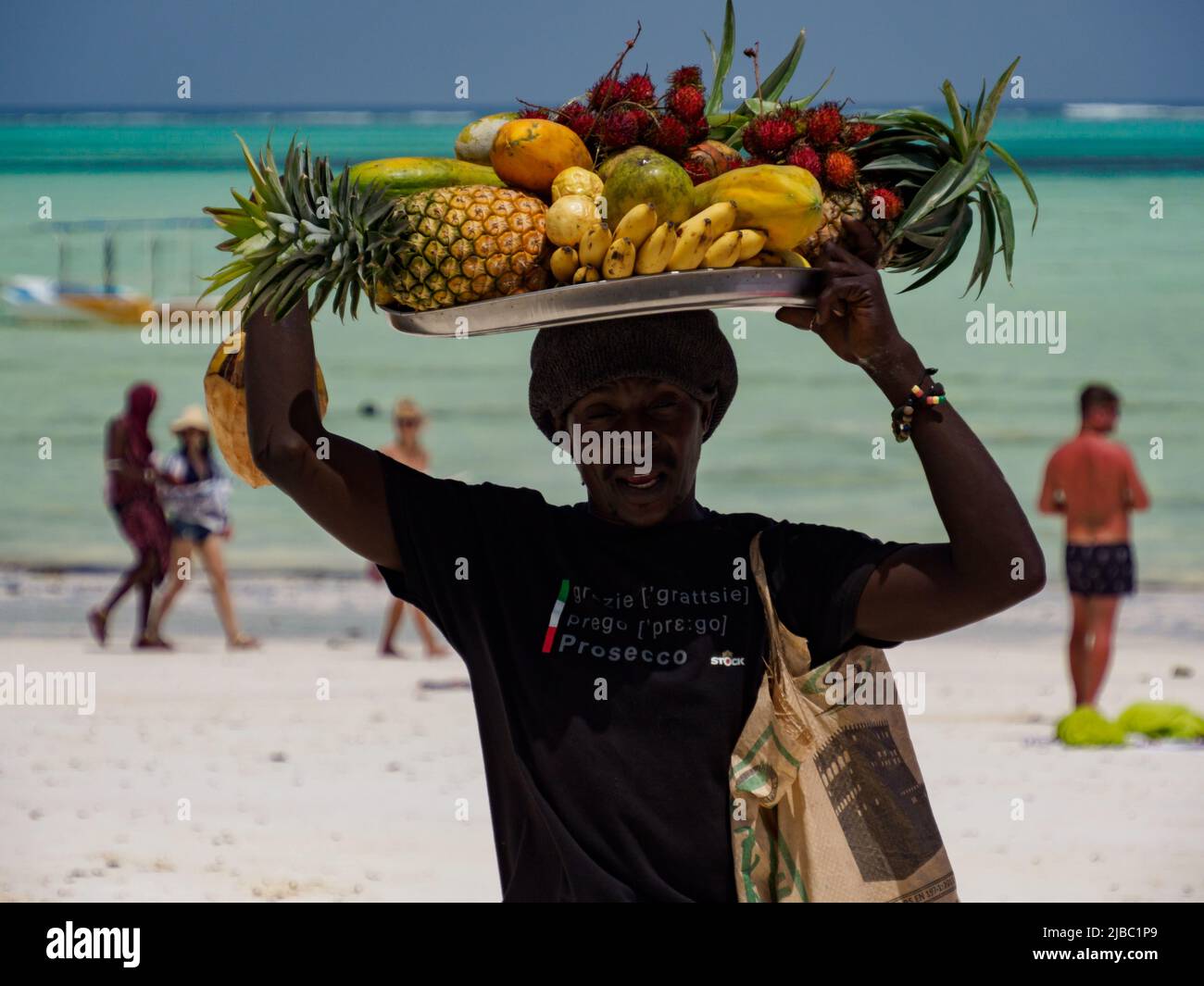 Paje, Zanzibar, Tanzania - January 202: African man with a tray full of exotic fruits on his head. Turquoise ocean water in the background. Africa Stock Photo