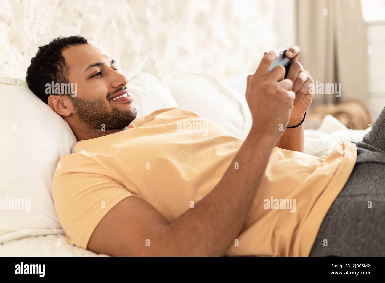 Cheerful Arabic Guy Using Cellphone Playing Game Lying At Home Stock Photo
