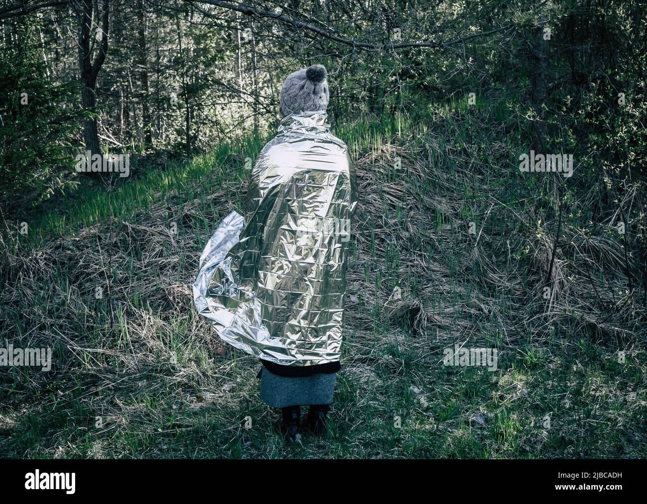 Woman person is lost and stand in wild forest in cold day, using first aid emergency blanket to prevent hypothermia and body heat loss. Stock Photo