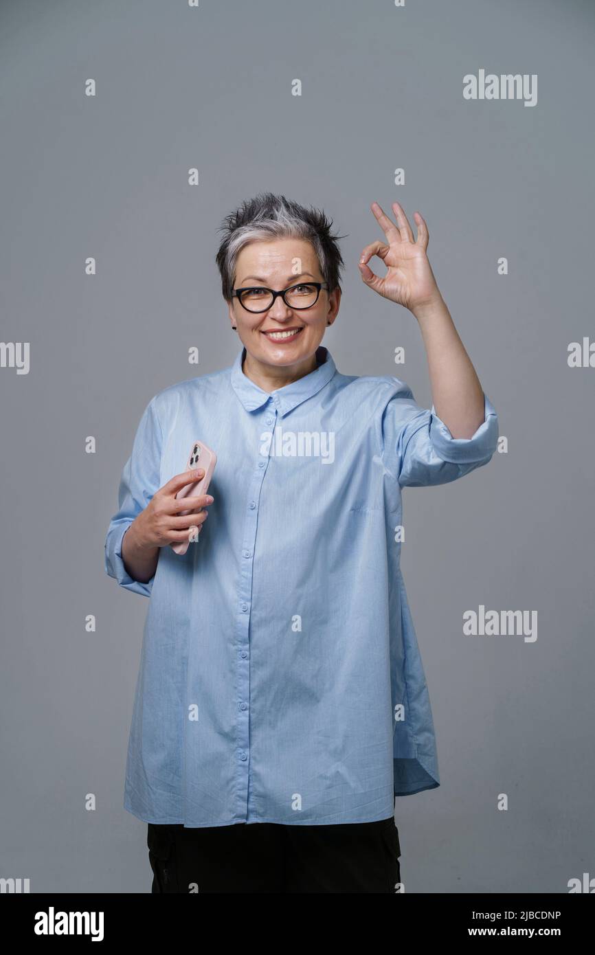 Gesturing OK sign grey haired mature businesswoman with smartphone in hand looking at camera wearing blue shirt. Pretty woman in blue shirt isolated on white. Toned image. Stock Photo