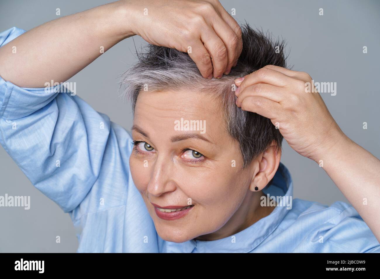 Pretty woman in 50s checking her hair or skull skin in mirror. Beautiful grey haired woman dealing with dandruff problem checking in mirror. Healthy hair, hair loss concept.  Stock Photo