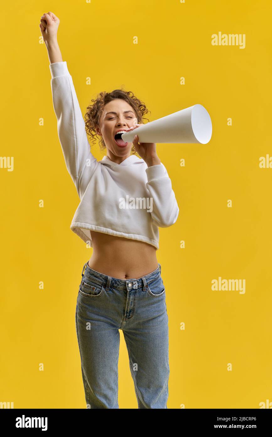Young woman shouting into megaphone, while holding one hand raised in studio. Front view of curly girl holding loudspeaker, announcing, isolated on orange background, copy space. Concept of announce. Stock Photo