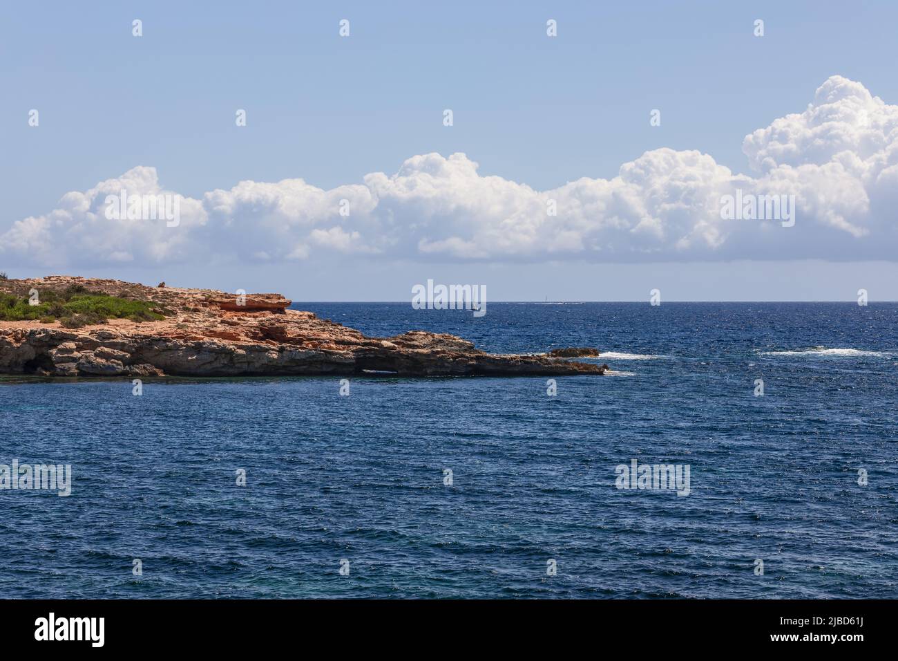 Small waves in bay bordered by flat rock gently descending to the sea clear sky with white cumulus low clouds, Ibiza, Balearic Islands, Spain Stock Photo