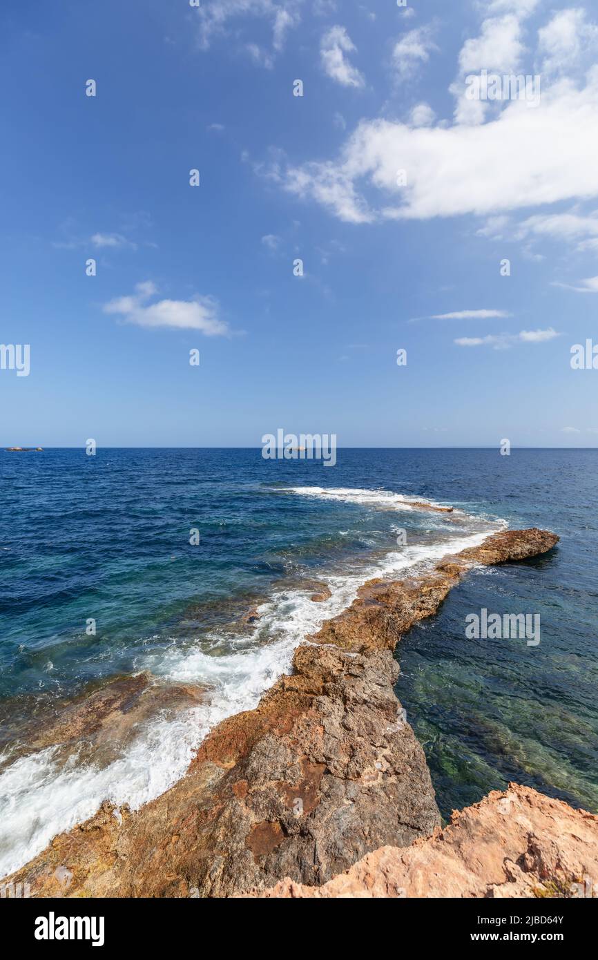 Vertical shot: narrow strip of rock gently descends into sea waters and is lost somewhere in distance, leaving foam from sea waves, Ibiza, Balearic Is Stock Photo