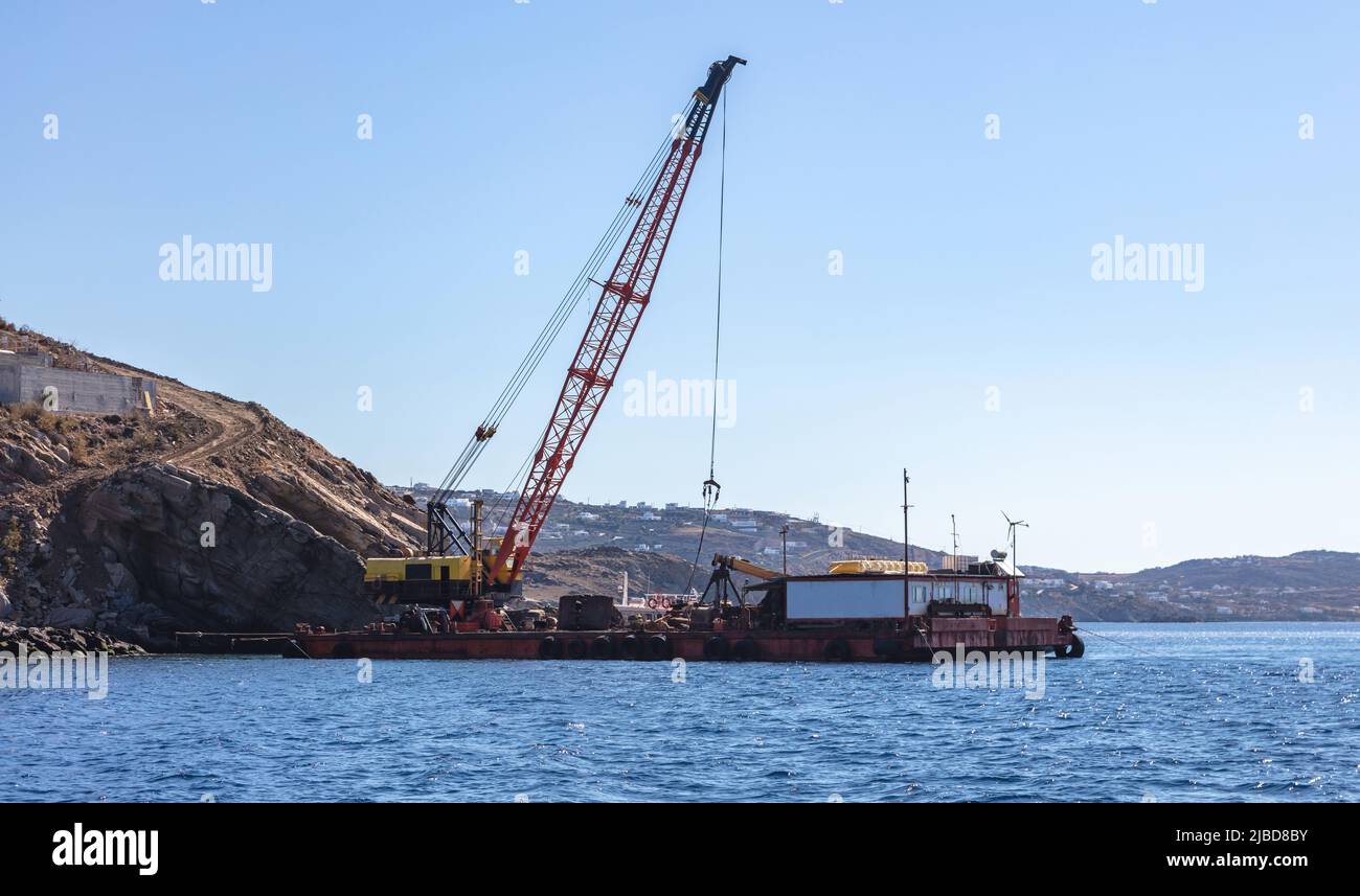 Marine works near Mykonos island port. Cyclades Greece. Construction machine, crane and barge on site, blue sky, rippled Aegean Sea Stock Photo