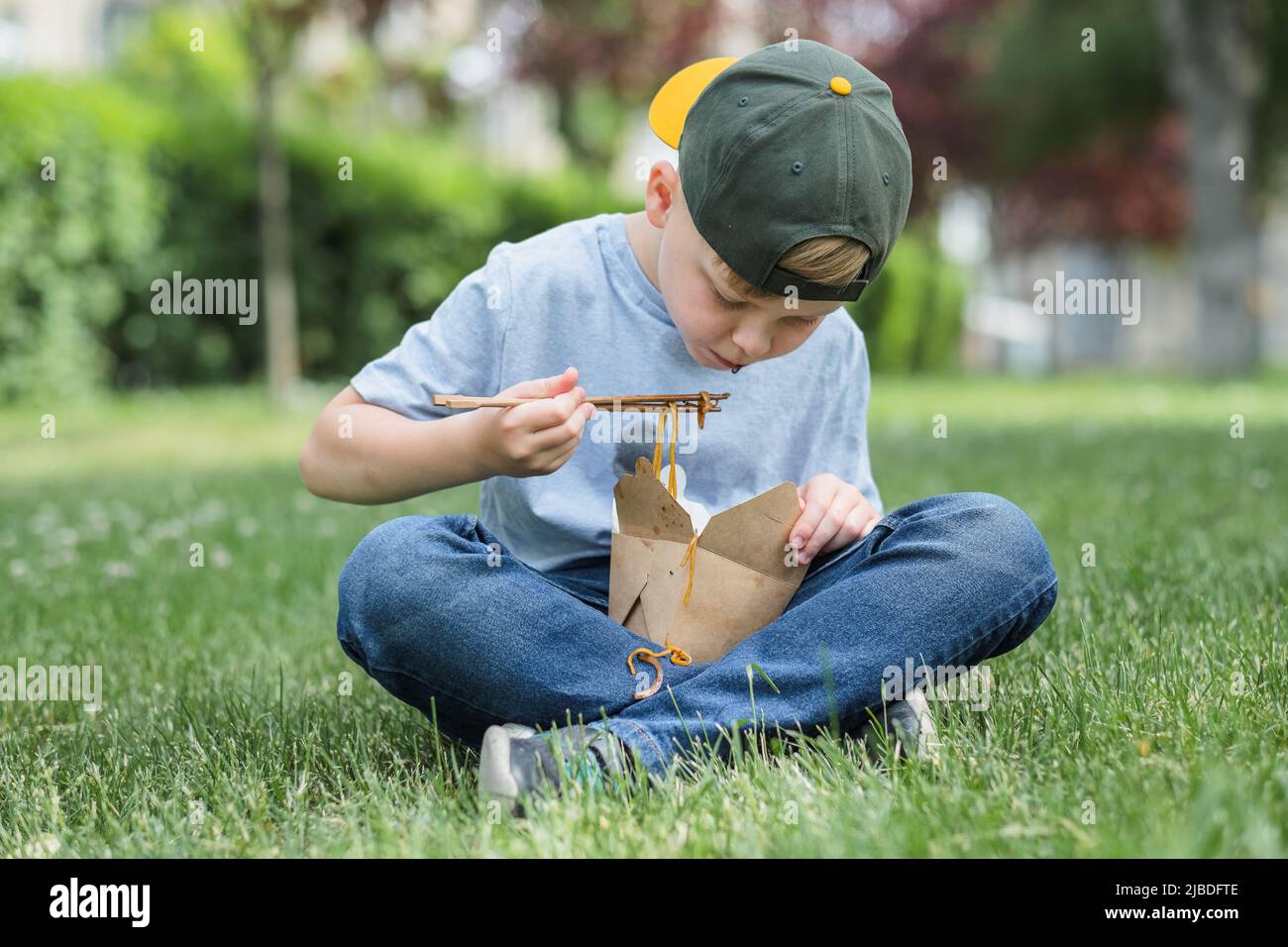 Child eating street food sitting on the grass. Dirty stain of sauce on his t-shirt. Noodles fell on his clothes. outdoors Stock Photo