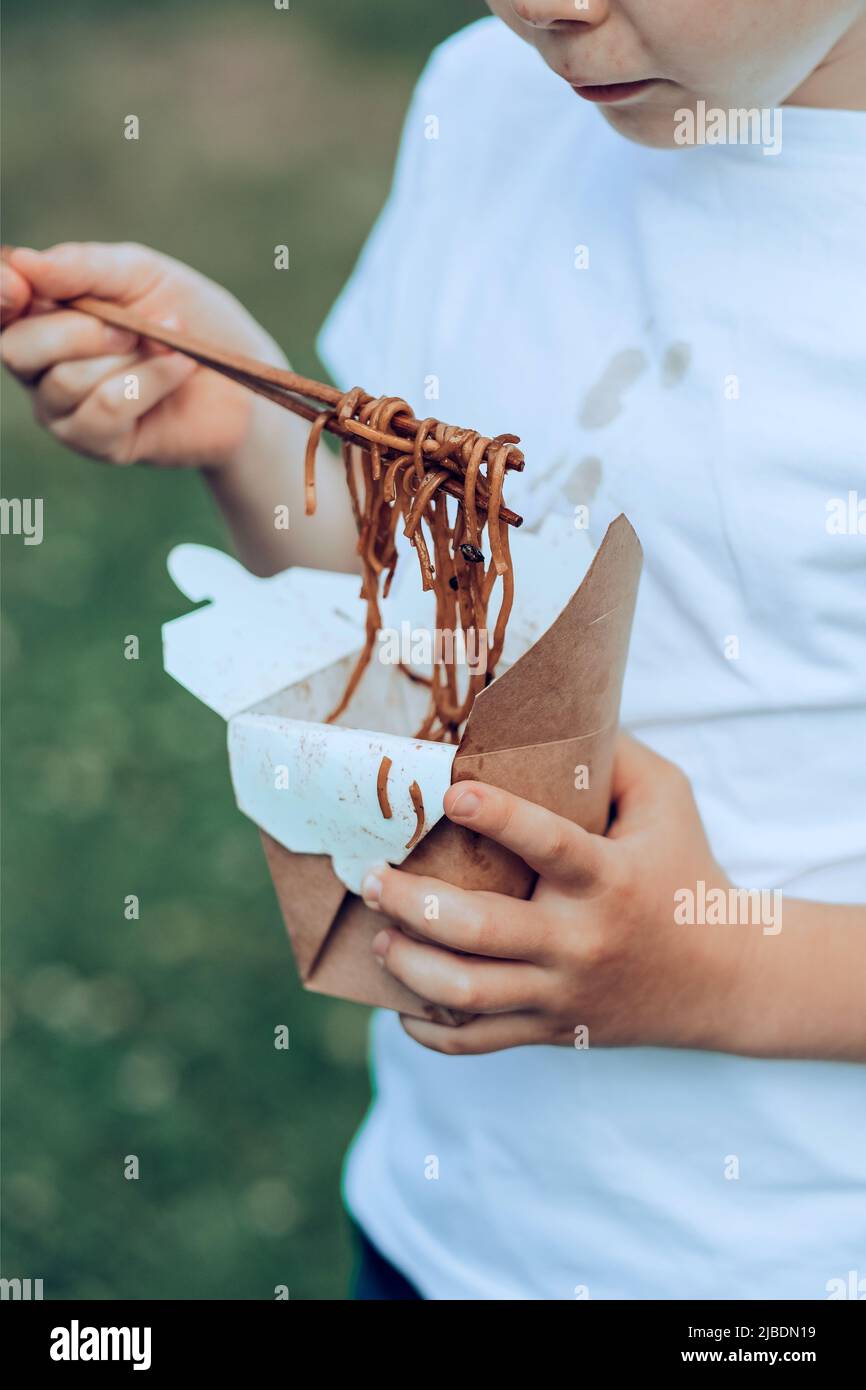 The child eating on the go soy sauce pan-fried noodles chopsticks. Dirty soy sauce stains on a white t-shirt. Takeaway food. outdoors Stock Photo