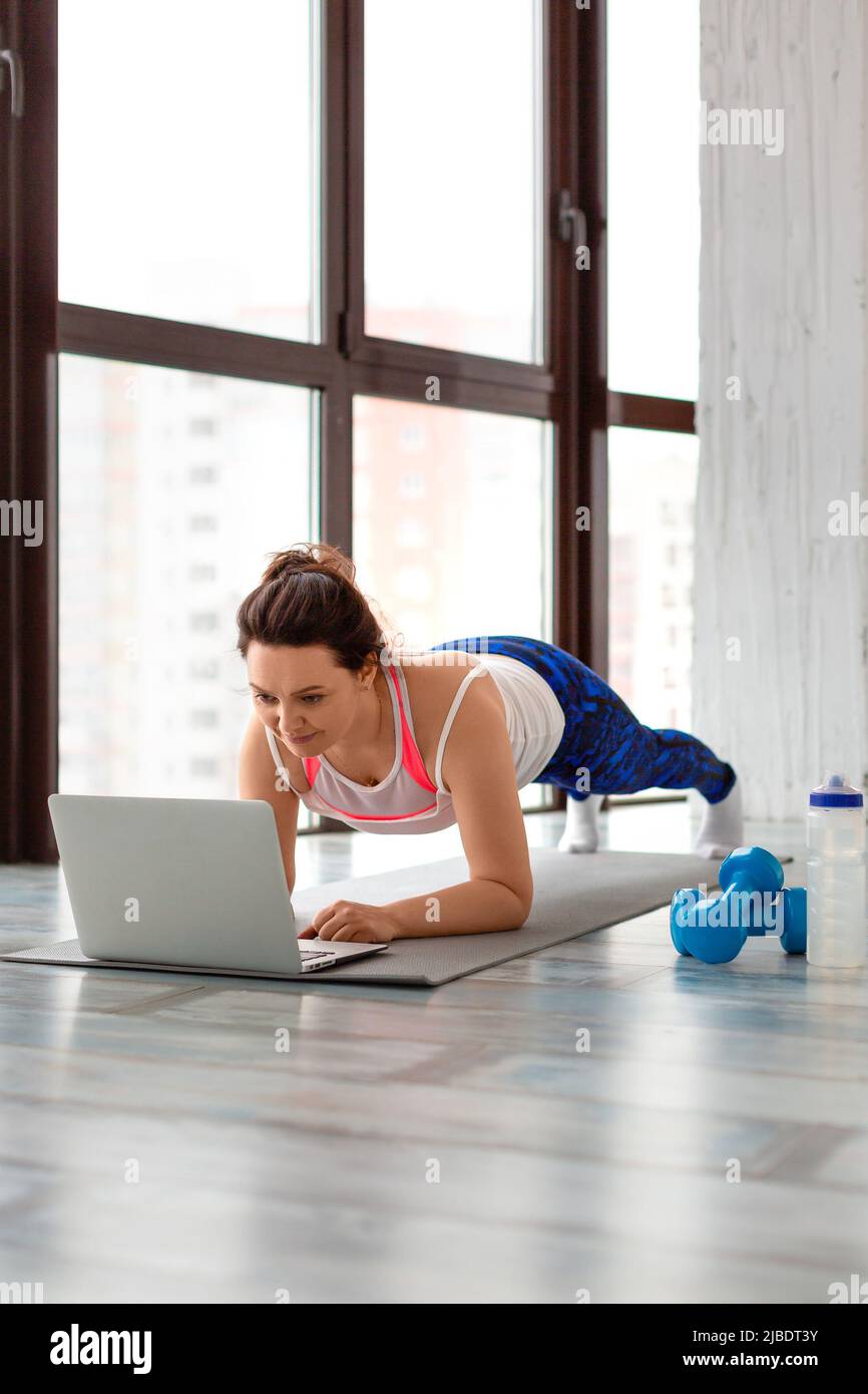 Middle-aged woman exercising at home online in front of a laptop monitor. In plank position. Healthy lifestyle concept. Vertical photo. Stock Photo