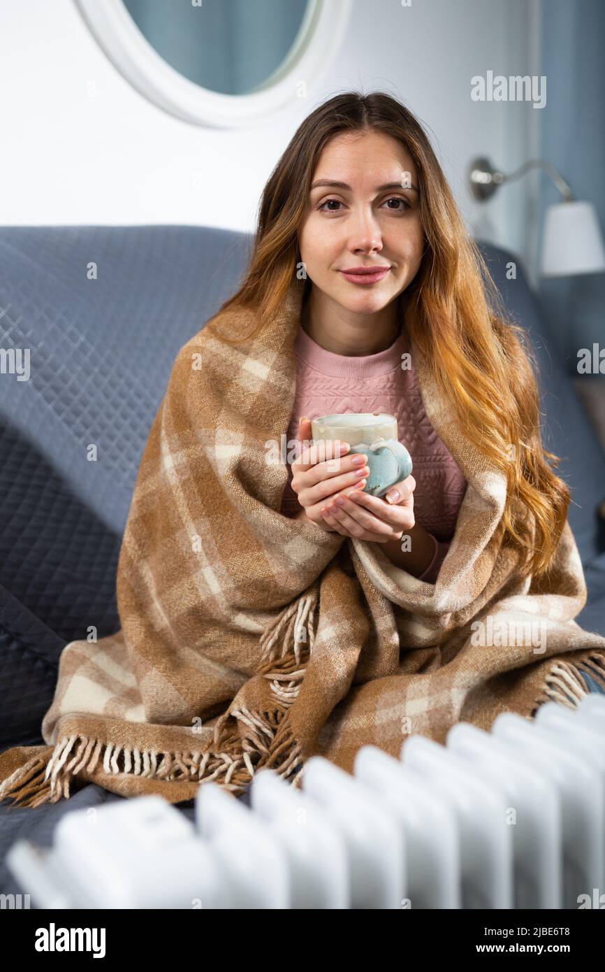 Woman with a blanket on his shoulders with cup near electric heater Stock Photo