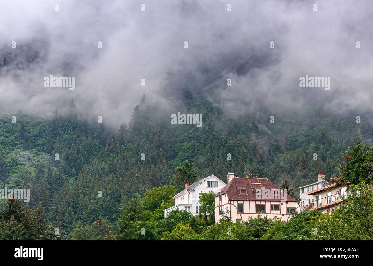 Juneau, Alaska, USA - July 19, 2011: Small group of larger houses peek out of pine forest on slopes of mountain, partly covered by low hanging gray cl Stock Photo
