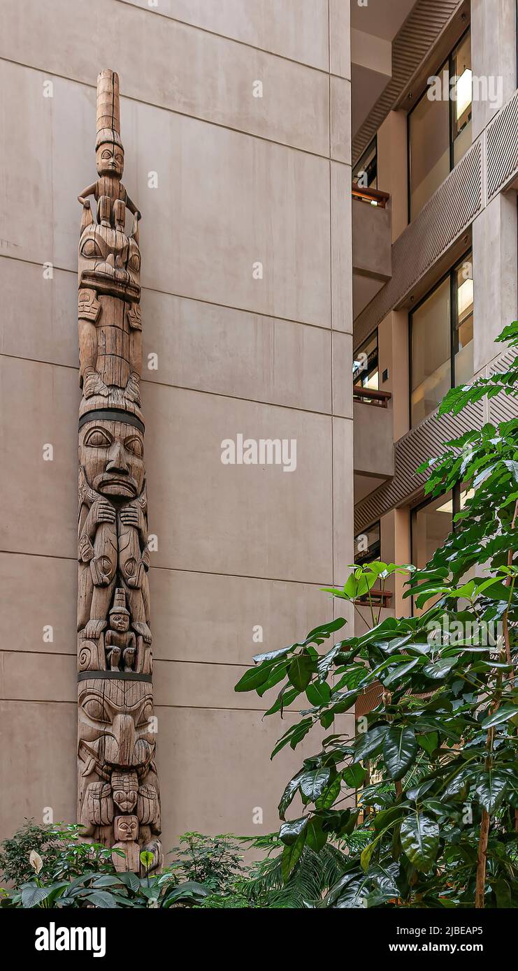 Juneau, Alaska, USA - July 19, 2011: Closeup of beige wood colored totem pole against beige wall of office building. Green foliage in front. Stock Photo