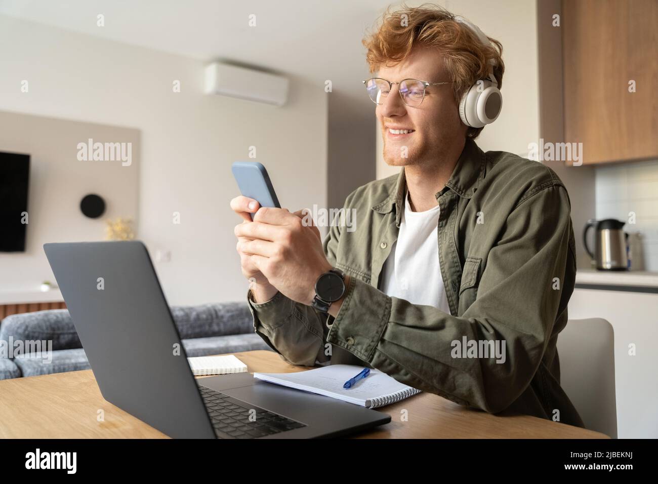 Young ginger man using smartphone while working online on laptop at home office Stock Photo