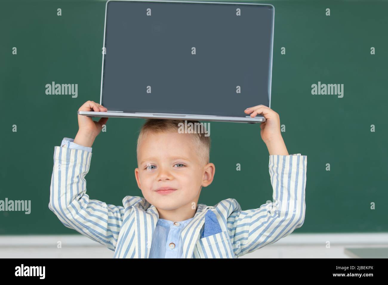 School child holding laptop on head using digital pc laptop in class. Portrait of funny pupil of primary school study programming. Stock Photo