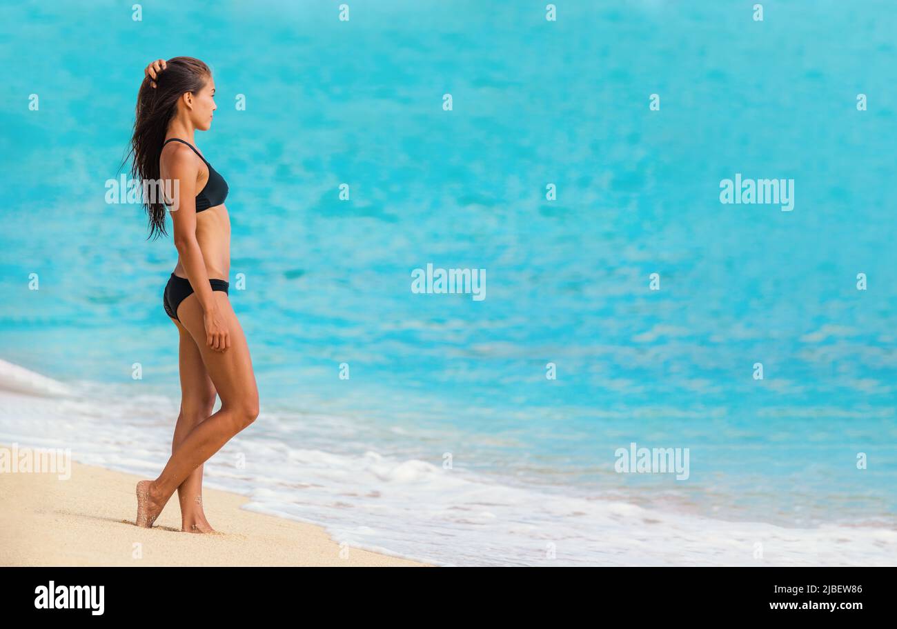 Asian bikini body woman going swimming in Caribbean ocean on beach vacation. Beautiful swimsuit model with lean legs and body profile wearing black Stock Photo