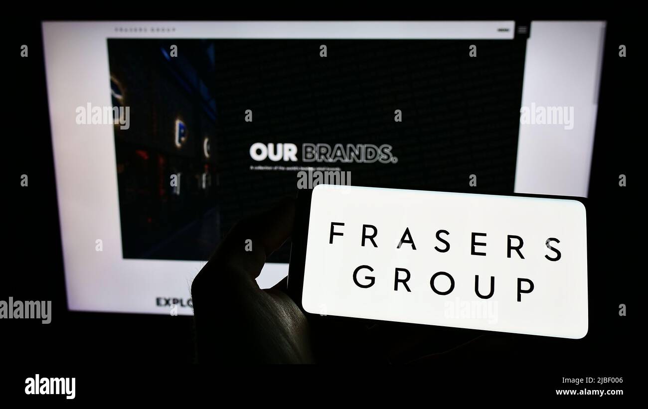Person holding mobile phone with logo of British retail company Frasers Group plc on screen in front of business webpage. Focus on phone display. Stock Photo