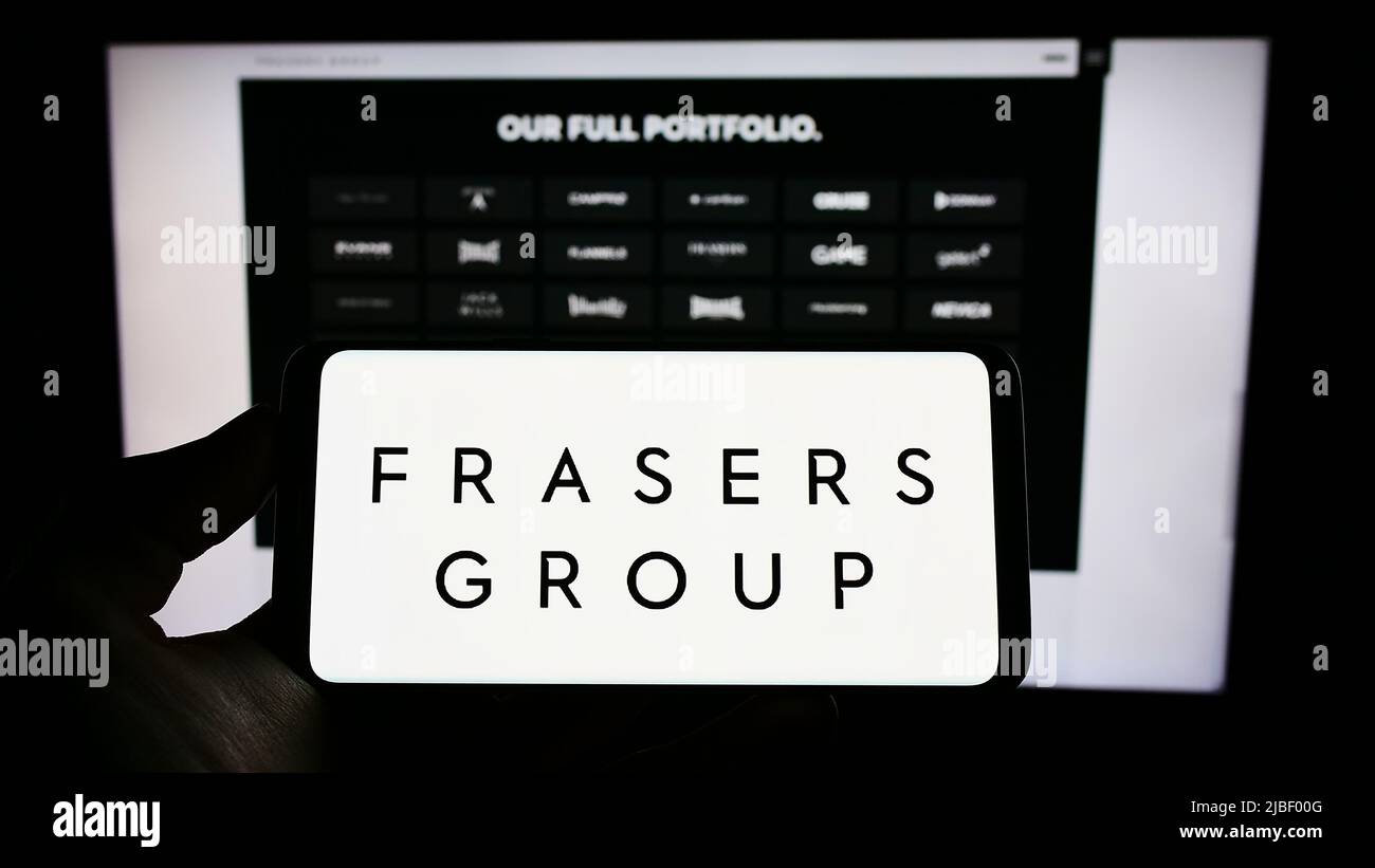 Person holding cellphone with logo of British retail company Frasers Group plc on screen in front of business webpage. Focus on phone display. Stock Photo