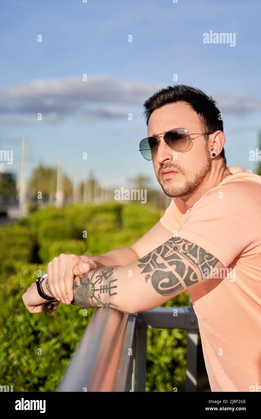 Side view of a serious young man, T-shirt and sunglasses, with a tattoo on his arm, standing on a bridge and looking away on a sunny day on a city stree Stock Photo