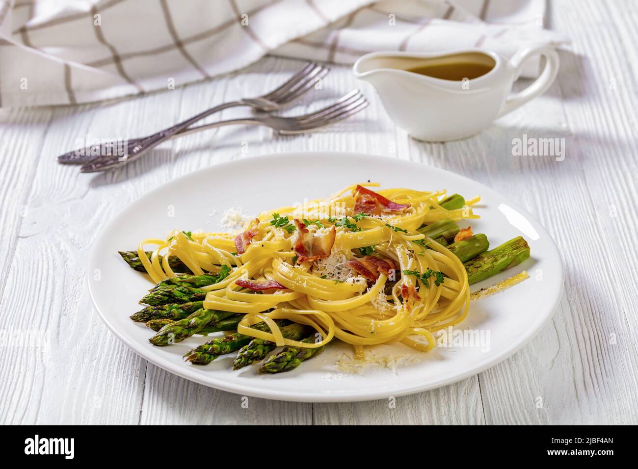 roasted asparagus spears with spaghetti sprinkled with shredded pecorino cheese, fried bacon pieces and parsley on white plate on white textured wood Stock Photo