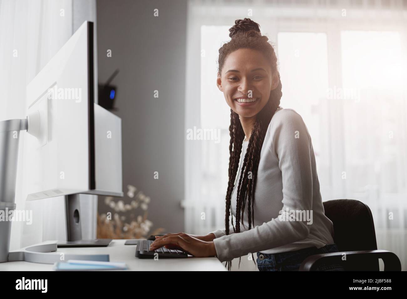 Portrait of pretty mixed-race young female worker sitting at desk in apartment room and smiling Stock Photo