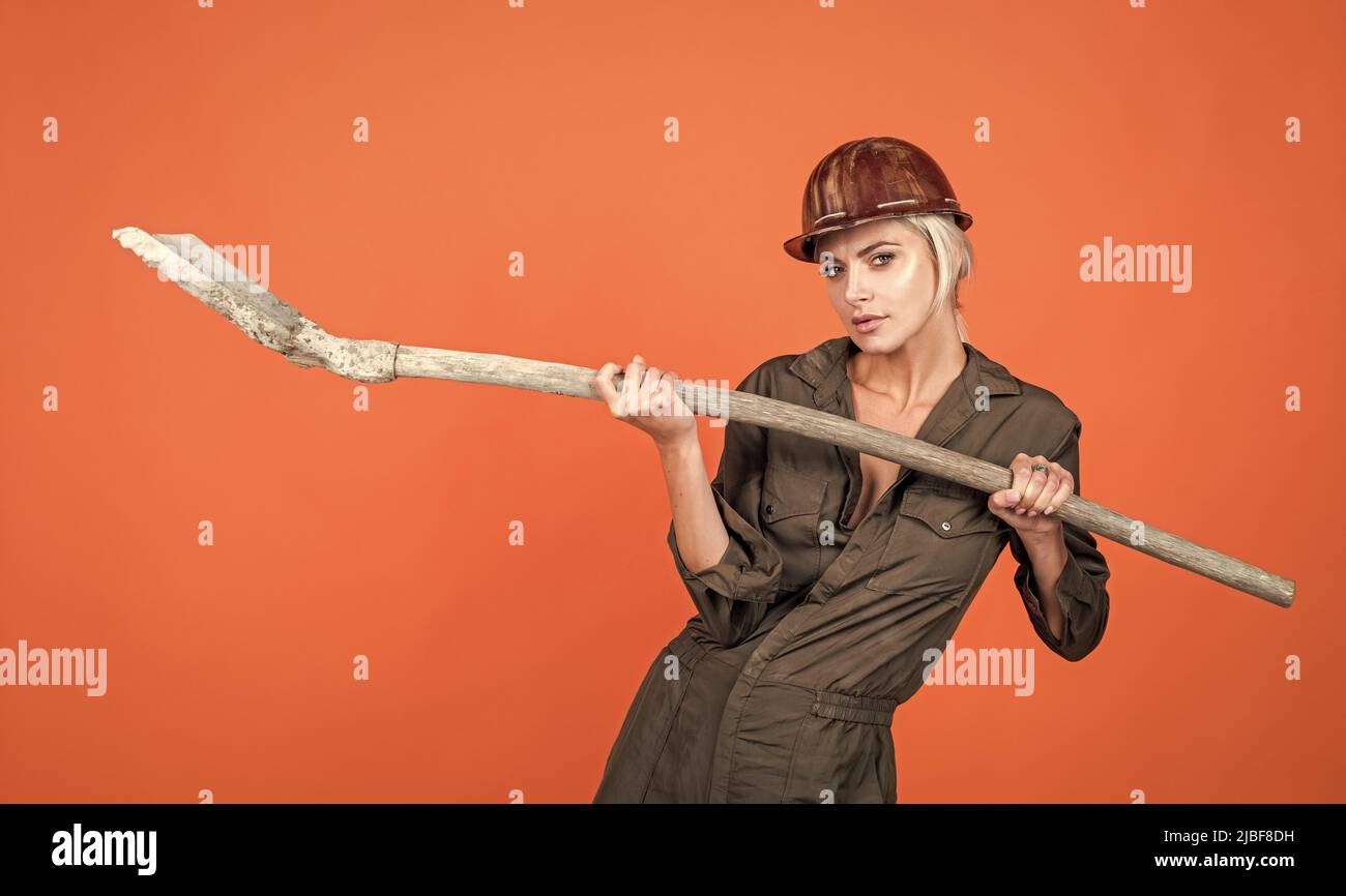 woman architect in protective helmet and boilersuit hold shovel on orange background, engineering Stock Photo