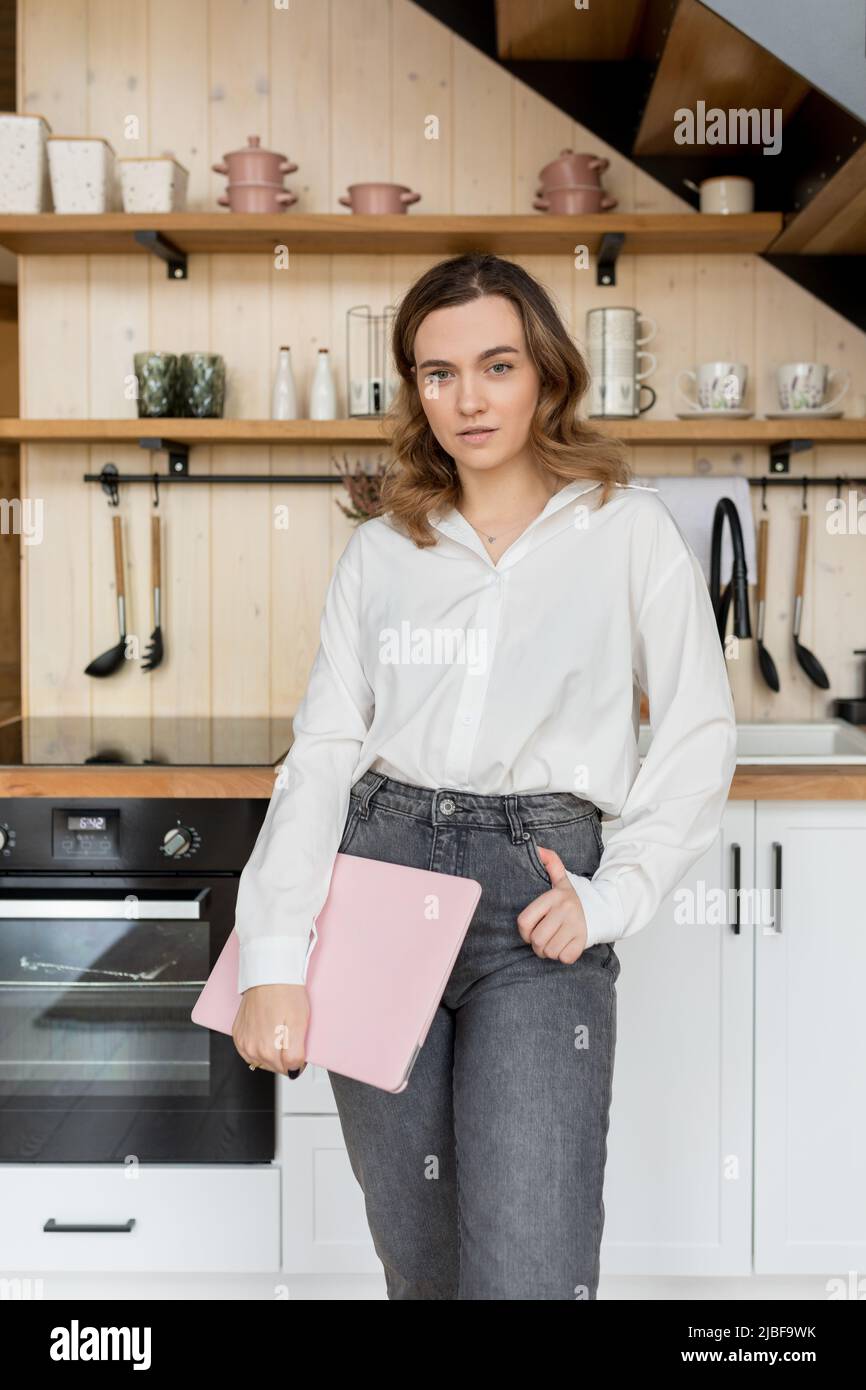 Interior designer. Charming brown-haired woman with long hair in white shirt and jeans stands against background of fragment of kitchen with laptop in Stock Photo