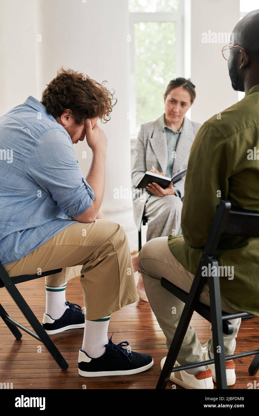 Fat young man touching his head while sitting in front of psychologist and describing his problem to her among other patients at session Stock Photo