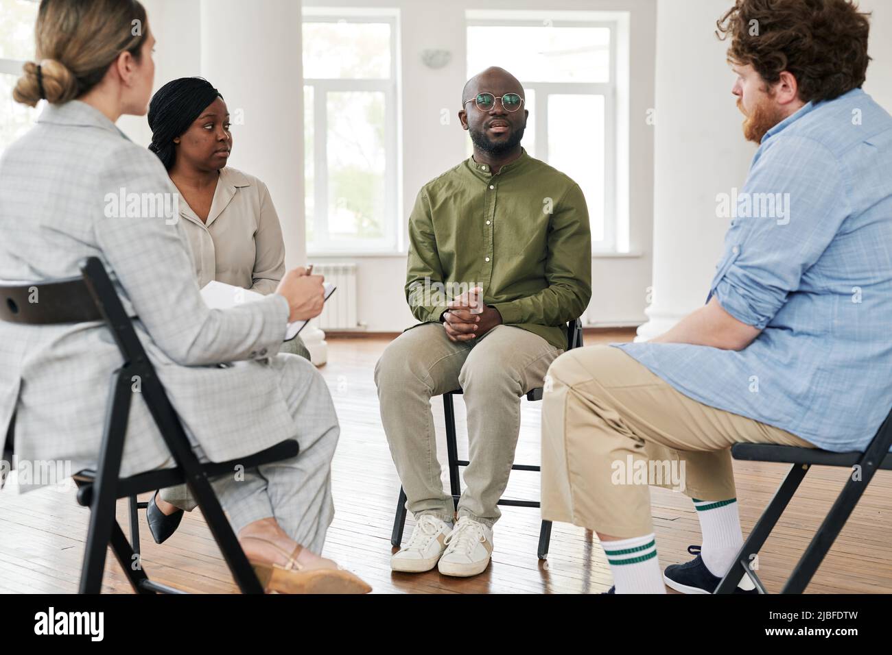 One of interracial patients describing his problem to psychologist and other people while sitting among them during session in office Stock Photo