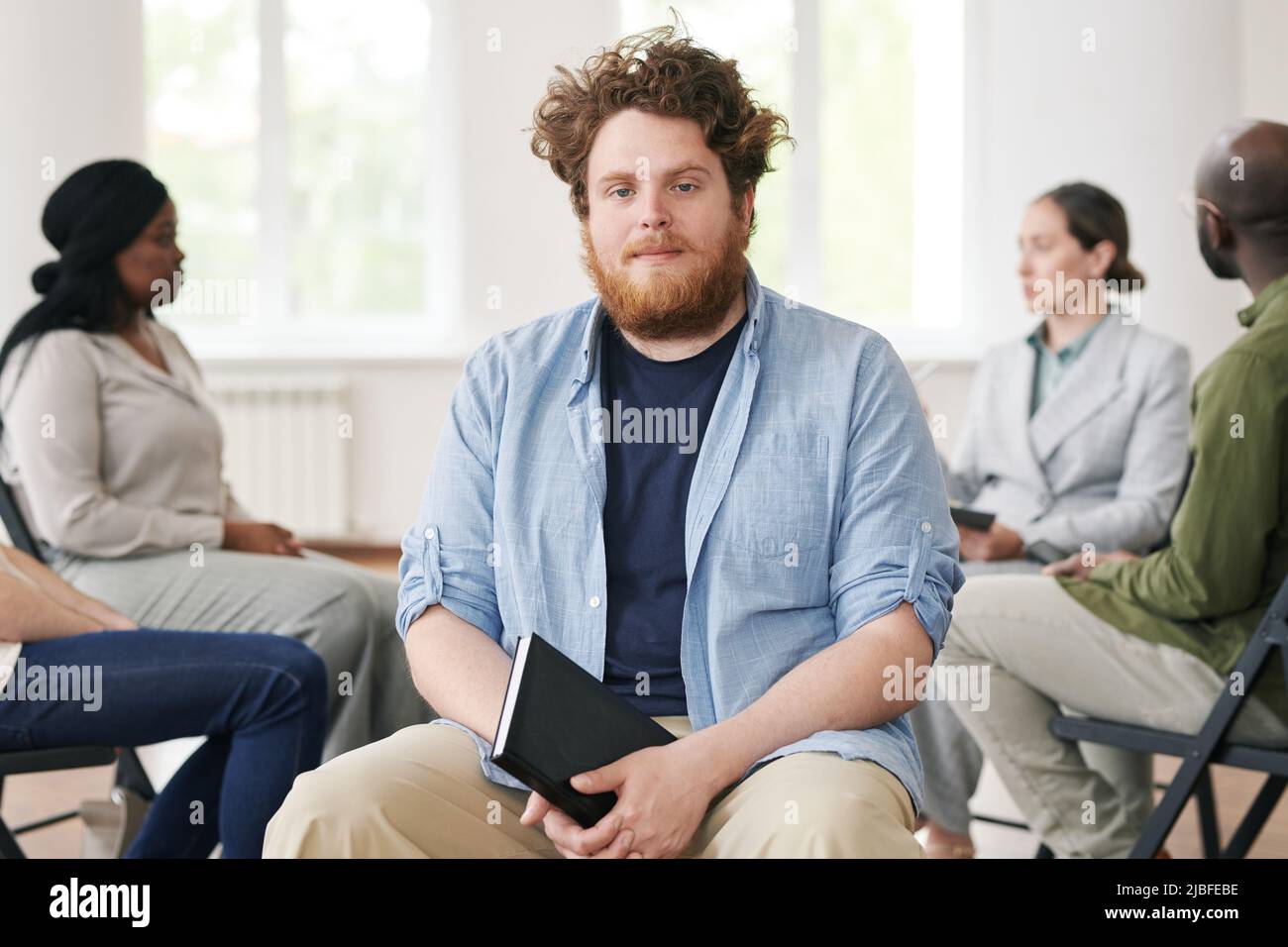 Young bearded man in casualwear holding notebook in black leather cover and looking at camera against group of people having session Stock Photo
