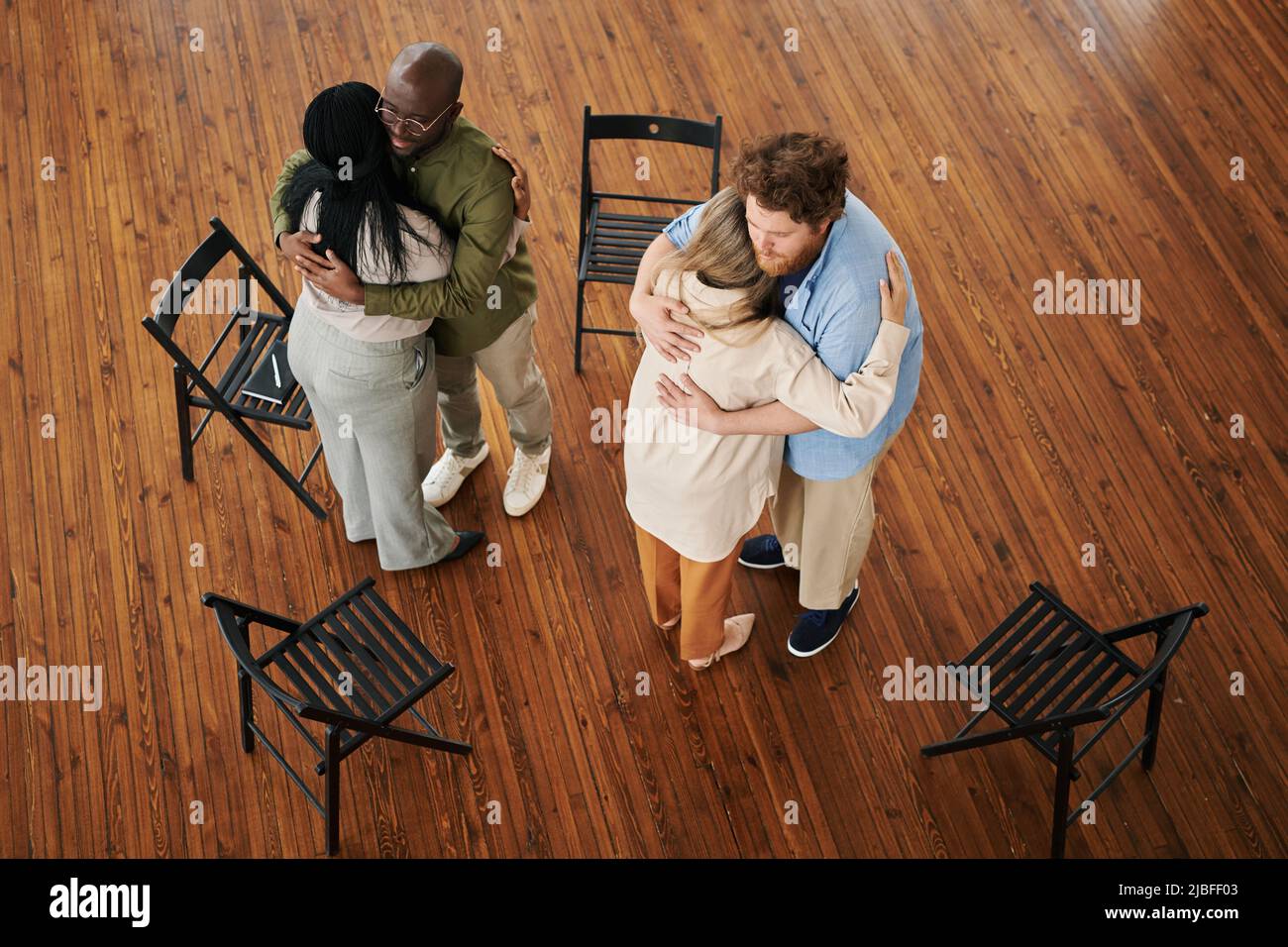 Two young interracial couples standing in embrace on the floor during psychological session while comforting each other Stock Photo