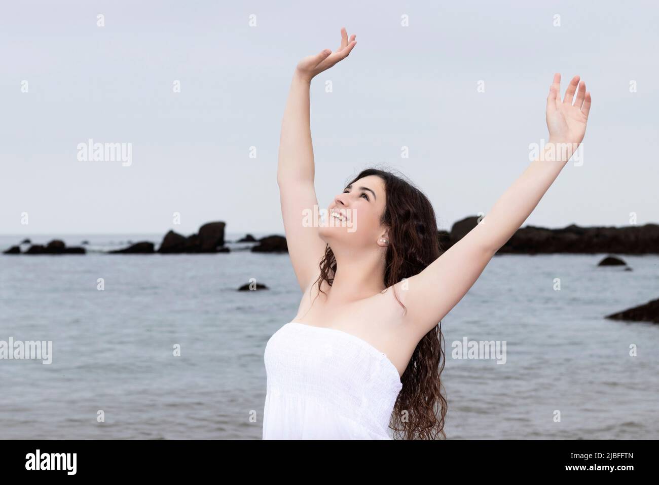 young brunette woman raising her arms on the beach in a white dress ...