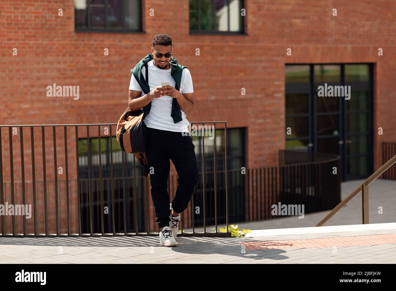 Trendy african american man surfing internet on cellphone, standing outdoor in city, texting on phone and smiling Stock Photo