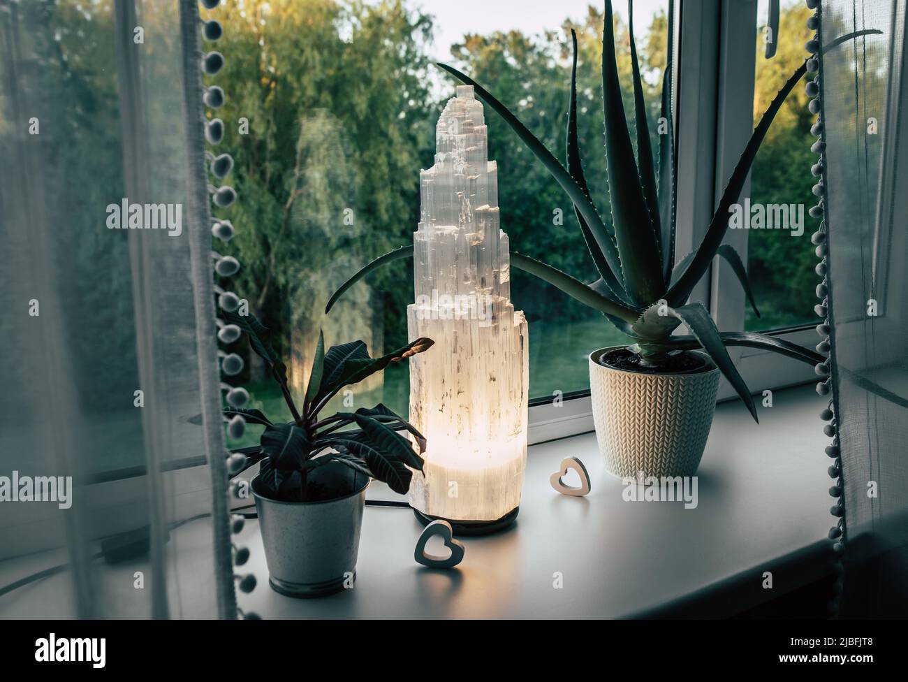 Rough big selenite crystal tower pole lamp illuminated in home on window sill, summer forest on background, spiritual home decor accent and ambient mo Stock Photo