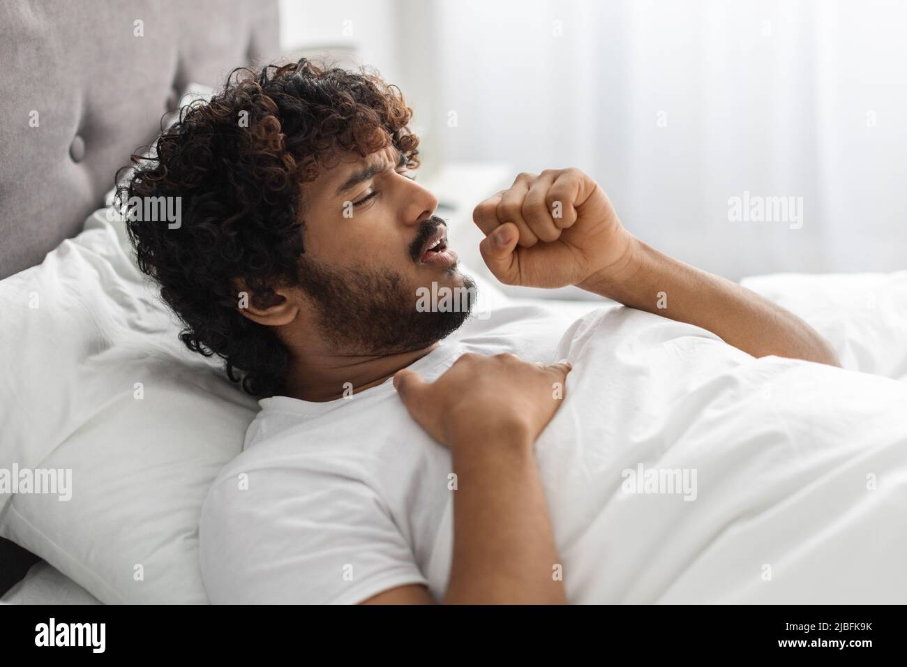 Sick indian man lying in bed, coughing and touching chest Stock Photo